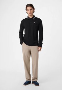 Svart långärmad pikétröja med vit krage och en liten logotypmärke, kombinerad med beige byxor och marinblå skor.