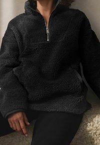 Personne portant un pull sherpa polaire noir zippé assise sur une surface claire et texturée.