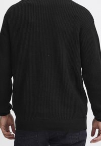 Osoba oblečená v černém dlouhém pleteném svetru a tmavých džínách, zobrazená zezadu s rukama volně podél těla.