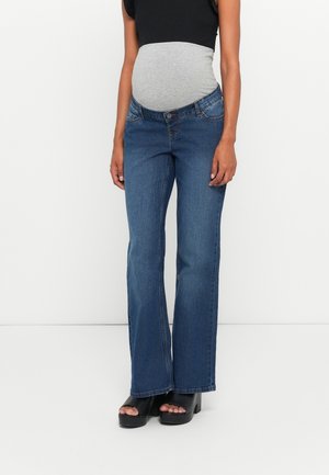 Personne enceinte portant un jean taille haute bleu avec une ceinture élastique grise et des chaussures à plateforme noires à bout ouvert.