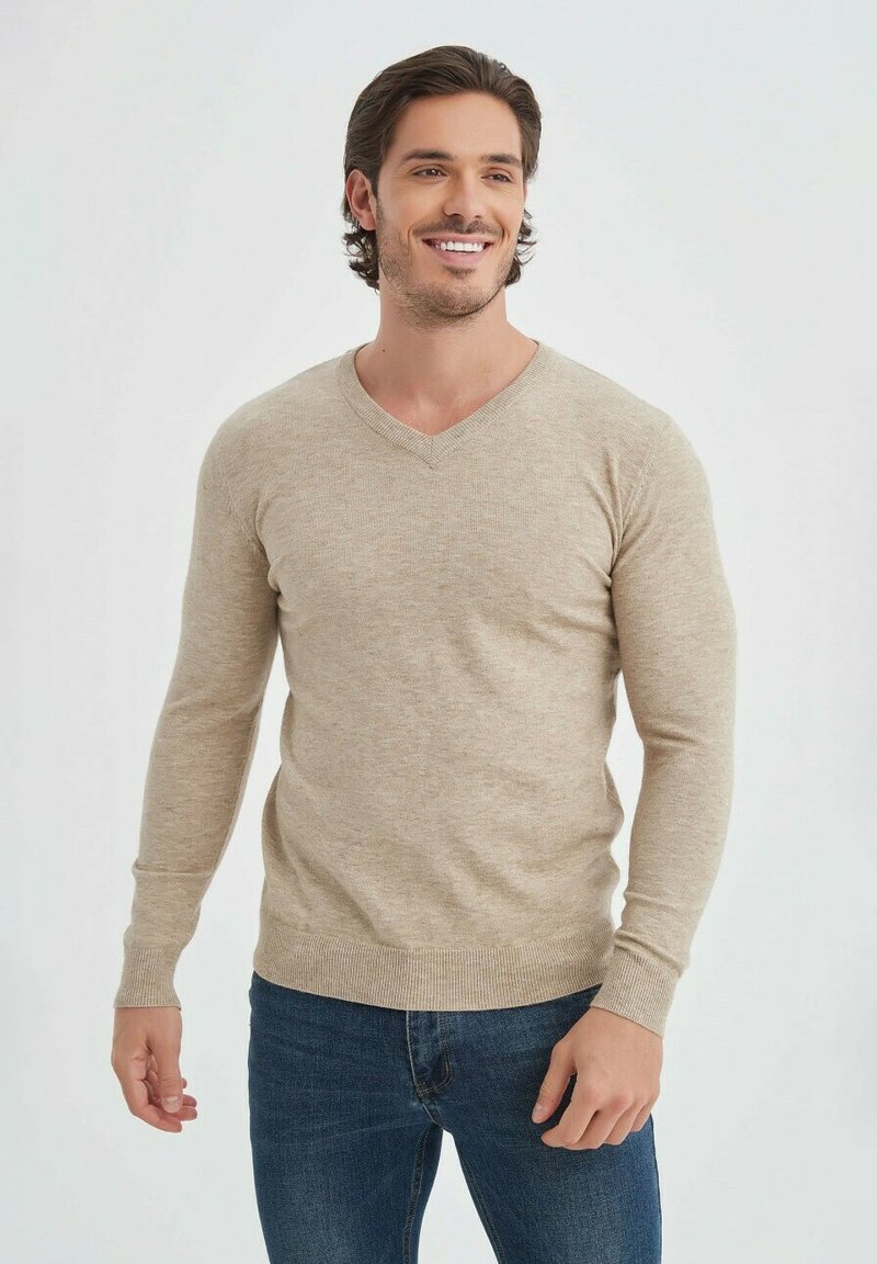 Pull en V beige à manches longues, présentant un design en tricot texturé. Porté sur un jean bleu foncé, exposé sur un fond neutre.