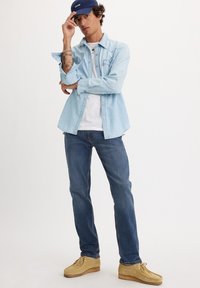 Helle blaue Jeanshemd mit Knopfleiste und Brusttasche, weißes T-Shirt darunter, dunkelblaue Jeans und beigefarbene Mokassinschuhe. Blaue Kappe.