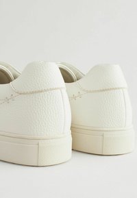 White leather sneakers with a textured finish, featuring a low profile and rubber soles, are seen from the back. Subtle stitching details present.