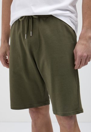 Uomo che indossa pantaloni corti con cordoncino verde oliva e una camicia bianca, in piedi con postura rilassata su uno sfondo semplice.