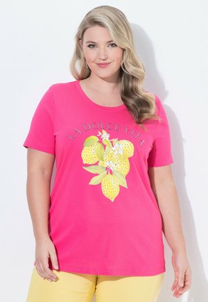 Vrouw met lang golvend blond haar die een felroze T-shirt draagt met de tekst "LA DOLCE VITA" en een citroengrafiek, gecombineerd met gele broek.