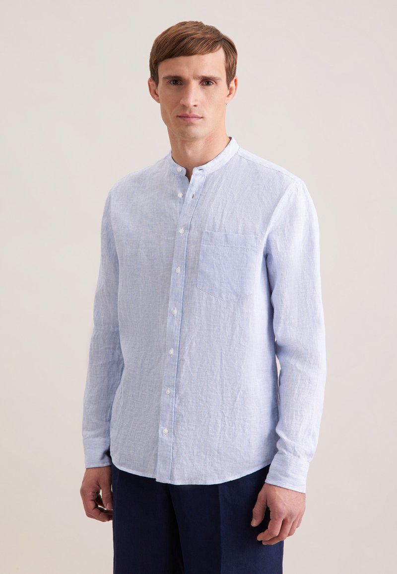 Jeune homme portant une chemise en lin bleu clair à col mao et un pantalon foncé, debout devant un fond beige uni.