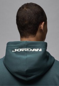 Sudadera de color verde azulado oscuro con un gran logo blanco de "JORDAN" en la parte posterior. Presenta una textura suave, costuras visibles y una capucha con cordón.
