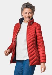 Veste matelassée rouge avec un col haut, fermeture éclair et doublure bleue contrastante. Présente des coutures horizontales et des poches zippées.