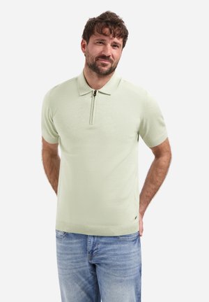 Man met kort bruin haar en baard, gekleed in een lichtgroen poloshirt met korte mouwen en een rits aan de voorkant, en lichtblauwe spijkerbroek, handen achter de rug.