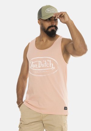 Homme avec barbe et anneau au nez portant un débardeur rose Von Dutch, un short cargo beige et une casquette olive Von Dutch ajustant la visière de la casquette.