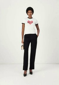 Žena u bijeloj majici s crvenim cvjetnim srcem, crnim obrubom, crnim hlačama i crnim štiklama, drži malu bež torbicu.