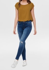 Olivgrön kortärmad topp, blå smala jeans med slitna detaljer och vita sneakers. Minimalistisk design med avslappnad passform.