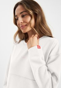 Witte sweatshirt met een geribde halslijn en boorden, voorzien van een roze geborduurd logo en een rode accent op de linker mouw. Zachte stofstructuur.