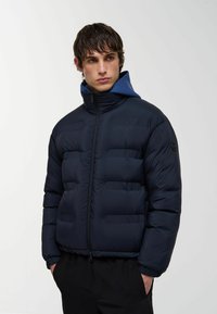Jachetă puffer de marină cu fermoar complet, guler înalt și design matlasat orizontal. Include o glugă interioară de un albastru contrastant și detalii cu logo.