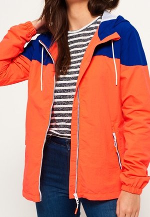 Młoda kobieta ubrana w pomarańczowo-niebieską kurtkę z kapturem zapinaną na zamek, na czarno-białej, paskowanej koszulce i niebieskich dżinsach.