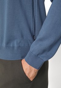 Modrý pletený svetr s žebrovanými manžetami a lemem, se zipovým detailem na boku. Materiál vypadá měkce a má jemnou texturu.