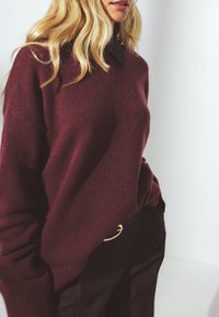 Burgundas krāsas adīta džemperis ar apkakles izgriezumu, brīva piegriezuma un ar ribotām piedurknēm. Kombinēts ar tumšām biksēm, kas izceļas ar zelta jostas akcentu.