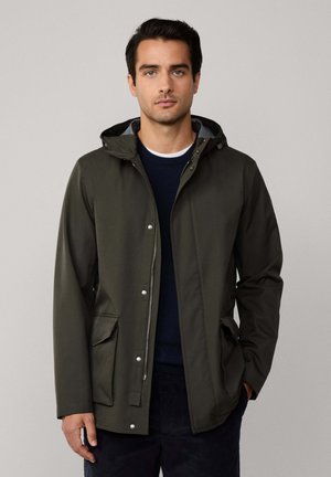 Jonge man in een olijfgroene hooded jack met klepzakken en een marineblauwe trui eronder, staande tegen een effen achtergrond.