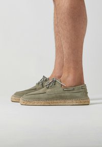 Espadrilles en daim vert avec une semelle plate en jute tissé, dotées de lacets gris et d'œillets en métal. Le design est décolleté et minimaliste.