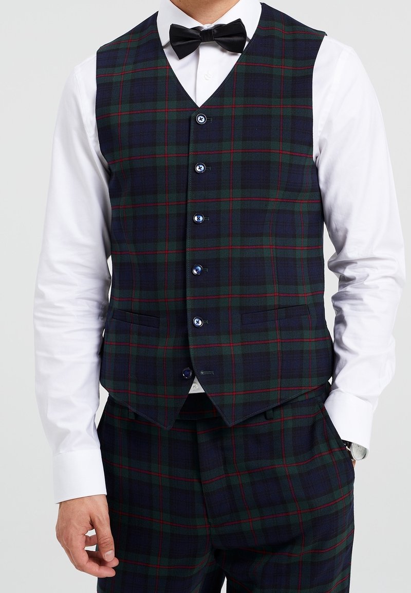 Chaleco de cuadros en azul marino, verde y rojo, con 5 cierres de botones azules, dobles bolsillos delanteros y un diseño de cuello en V. Llevado sobre una camisa blanca.