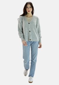 Grau gerippter Cardigan mit V-Ausschnitt, ausgestattet mit braunen Knöpfen; getragen über einem hellen Shirt, kombiniert mit blauen Jeans und weißen Sneakers.