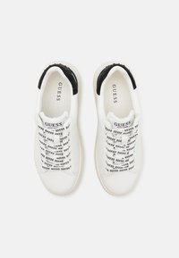 Sneakers bianchi con un tallone nero texturizzato, caratterizzate da un motivo di lacci con il testo ripetuto "GUESS" e una punta liscia e arrotondata.