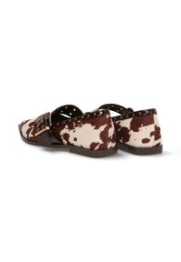 Zapatillas slip-on con estampado de vaca en tela marrón y crema, de perfil bajo y con una correa decorativa con hebilla. Suela plana y texturizada.