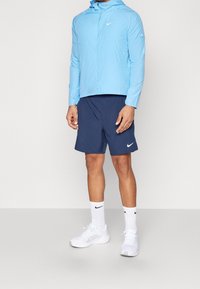 Veste légère bleu clair avec capuche, associée à un short bleu marine, des chaussures de sport blanches et des chaussettes blanches avec un logo noir.
