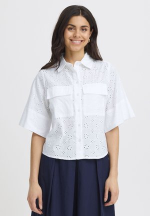 Tânără zâmbitoare, purtând o cămașă albă cu mânecă scurtă, decorată cu găurele și cu buzunare pe piept, și o fustă bleumarin, pe un fundal simplu.