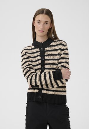 Femme aux cheveux bruns raides portant un cardigan boutonné à rayures noires et beiges ainsi qu'un pantalon noir, debout les bras croisés.