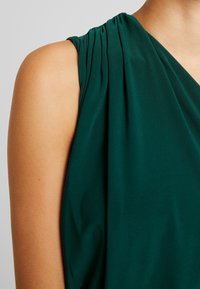 Šaty bez rukávů ve tmavě zelené barvě, s plisováním na rameni a hladkou texturou. Čisté linie s draped designem.