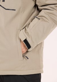 Beige jacka med justerbara ärmslut med kardborre, sidofickor och en smidig textur. Svarta dragkedjor syns.