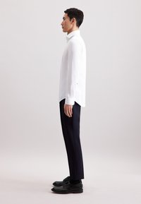 Camicia bianca a maniche lunghe con colletto strutturato, pantaloni neri su misura e scarpe nere. La camicia presenta una texture delicata e un orlo asimmetrico.