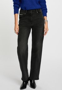 Jeans de pierna ancha negra de denim, con bolsillos frontales con acentos metálicos. Llevado con un suéter azul y botas de tacón negras.