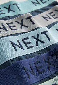Gros plan de quatre paires de sous-vêtements avec des ceintures élastiques en bleu, gris et marine, chacune comportant le mot « NEXT » en lettres grasses.