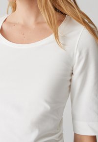 Femme portant un haut blanc à manches courtes avec un col arrondi, dévoilant une partie de son cou et ses cheveux blonds sur l’épaule.