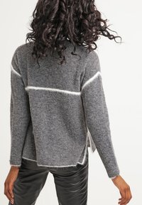 Pull tricoté gris avec des rayures blanches aux épaules, présentant une coupe ample, une texture douce et des fentes latérales. Accentuée par des poignets en côtes.