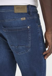 Temno modri jeans iz denima z zadnjim žepom, ki ima vzorec cik-cak šiva. Svetlo rjav oznaka na pasu nosi napis "BLEND." Gladka tekstura.