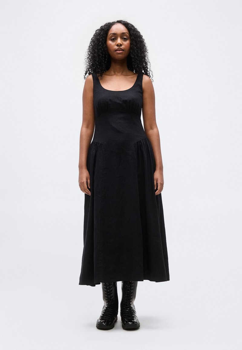 Γυναίκα με σγουρά μαλλιά φοράει μαύρο μίντι φόρεμα χωρίς μανίκια με στρογγυλή λαιμόκοψη και μαύρα μπότες με κορδόνια, στέκεται μπροστά σε απλό φόντο.