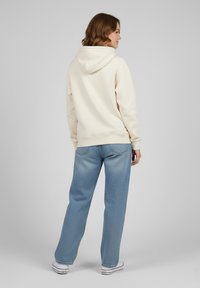 Felpa color crema con cappuccio regolabile, abbinata a jeans azzurri chiari e larghi con dettagli di texture sottili e classiche sneaker bianche.