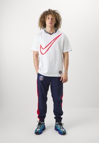 Camiseta blanca con un gran logo rojo de Nike, pantalones deportivos azul marino con franjas rojas y el logo del Paris Saint-Germain, y zapatillas deportivas azul-negro.