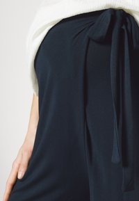 Námorně modrý oděv s měkkou texturou, s vázáním v pase, splývavým střihem a překryvem z bílé, texturované látky.