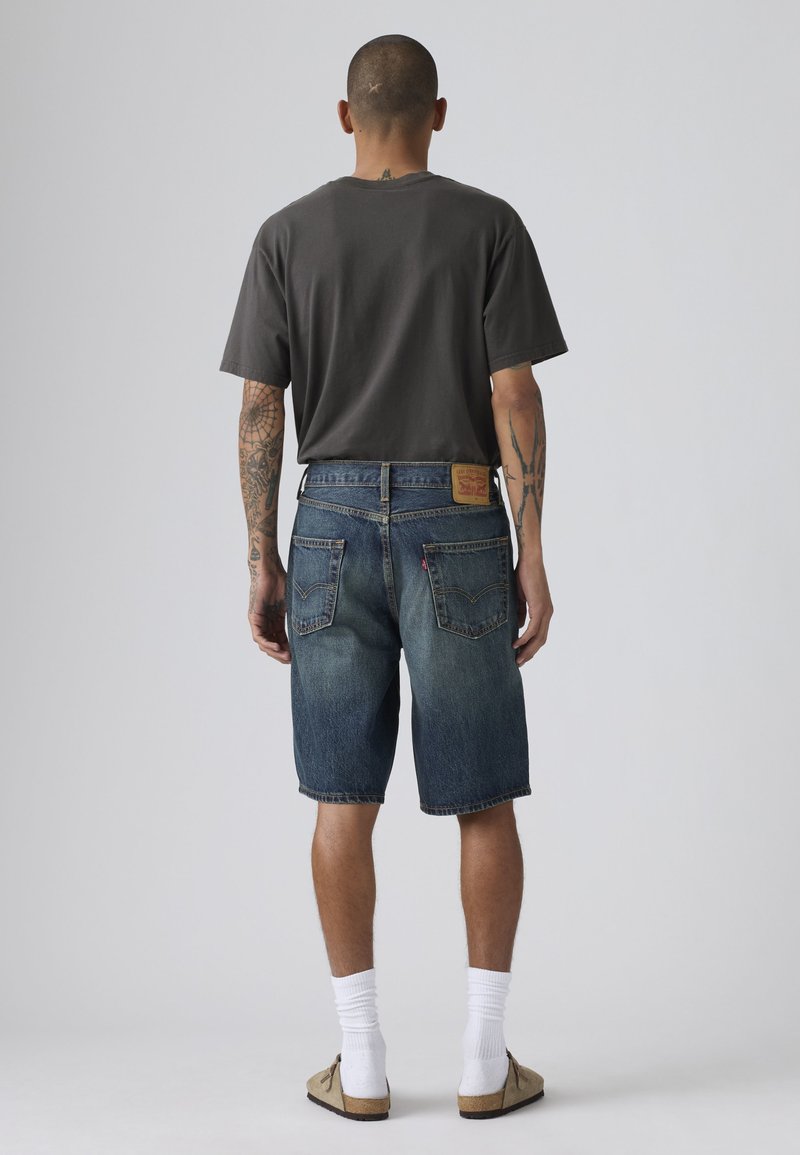 Homme tatoué portant un t-shirt gris foncé, un short en denim bleu, des chaussettes blanches et des chaussures beiges, debout de dos contre un fond uni.