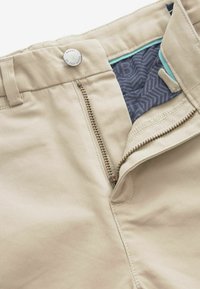 Pantaloni in cotone beige con zip, chiusura a bottone e fodera in tessuto blu a fantasia. Presentano cuciture visibili e una texture liscia.