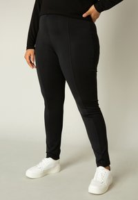 Zwarte leggings van gladde stof met een aansluitend ontwerp. Heeft zijn naden aan de zijkanten en een hoge tailleband. Draagt met witte sneakers.