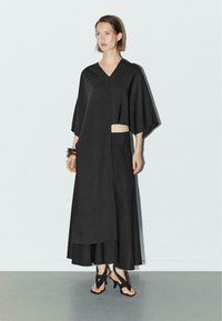 Kvinde i sort asymmetrisk V-hals top og vide bukser, iført hæltonder og armbånd, stående op ad en hvid væg.