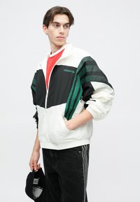 Balta un melna sporta jakas ar zaļām svītrām, ar rāvējslēdzēju un ribotām aprocēm. Modelis valkā sarkanu kreklu un tur melnu cepuri.