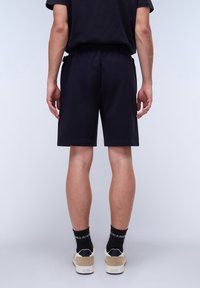 Shorts de sport noirs en tissu doux, taille élastique, longueur au-dessus du genou, associés à des baskets claires et des chaussettes noires mi-mollet.