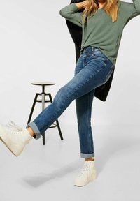 Blå denimjeans med en figurnära design och upprullade ärmslut. Kombinerat med en grön långärmad topp och vita hög-top sneakers. Minimalistisk bakgrund.