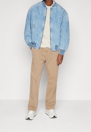 Personne portant une veste bomber en denim bleu clair, un pull crème, un pantalon beige avec des poches et des baskets blanches, debout devant un fond blanc.